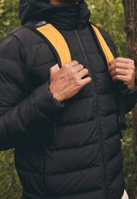 Musta puhvotakki vetoketjulla, jossa on tikkikuvio. Keltaiset olkaimet ja logo kauluksessa korostavat muotoilua.