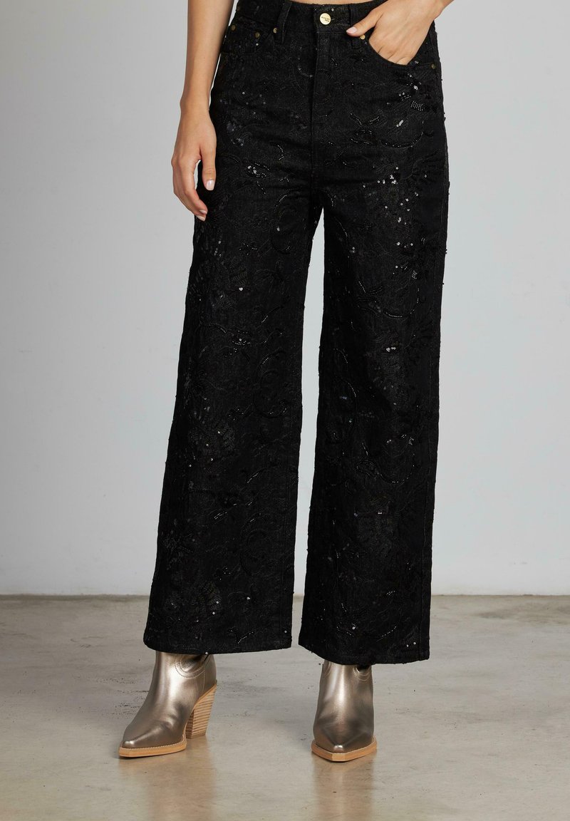 Pantaloni neri a gamba larga realizzati in tessuto testurizzato con motivi floreali, caratterizzati da sottili paillettes e vita alta, abbinati a stivali metallici alla caviglia.