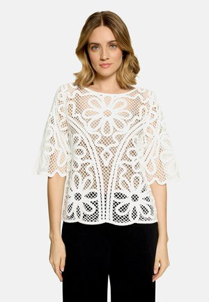 Femme portant un chemisier en dentelle florale blanche à manches larges et un pantalon noir, debout devant un fond clair uni.