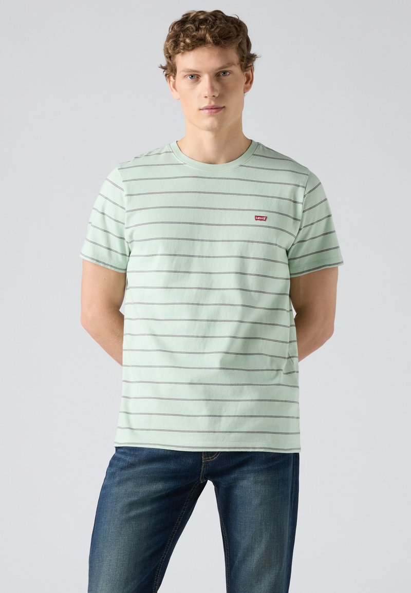Jeune homme aux cheveux bouclés portant un t-shirt à rayures vert clair et un jean bleu sur un fond gris uni.