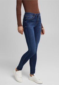 Jeans ajustados de denim azul oscuro, con un diseño clásico de cinco bolsillos, costuras sutiles y una cintura de altura media, combinados con zapatillas blancas.