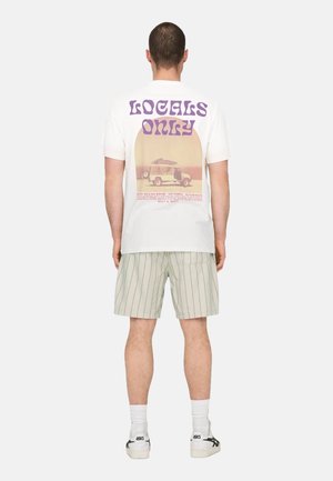 Witte t-shirt met de tekst "LOCALS ONLY" in paars en een vintage auto graphic. Gecombineerd met gestreepte shorts en witte sneakers.