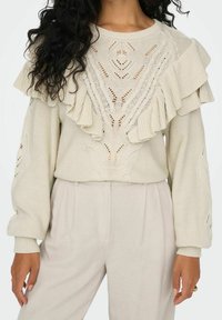 Beżowy dzianinowy sweter z falbaniastym dekoltem i skomplikowanymi wzorami ażurowymi, zestawiony z jasnobeżowymi spodniami o wysokim stanie.