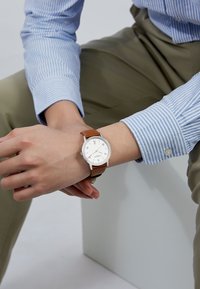 Montre argentée avec un cadran blanc et un bracelet en cuir marron, présentant un design minimaliste. Portée au poignet avec une chemise à rayures bleues.