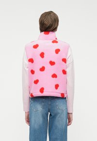 Άτομο ντυμένο με μπλε τζιν και ροζ φλις γιλέκο με κόκκινα σχέδια καρδιάς, που στέκεται με την πλάτη προς τον θεατή σε λευκό φόντο.