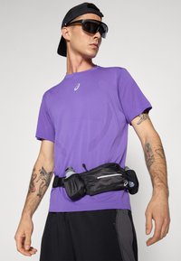 Lila Sportshirt, schwarzer Laufgürtel mit Wasserflaschen, sichtbarer tätowierter Arm und Sonnenbrille; lässiges Sportbekleidungsdesign und Zubehör.