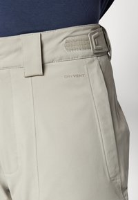 Beige broek van gladde stof met een textuurband, zijzakken en een "DRY VENT" label nabij het heupgebied.