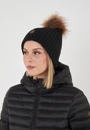 Donna che indossa una giacca nera trapuntata e un cappello di maglia nero con un pompon in pelliccia sintetica marrone, che guarda leggermente verso sinistra.