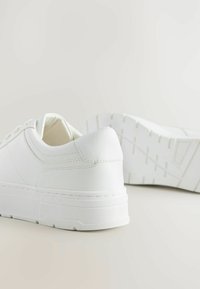 Sneakers bianchi realizzati in materiale sintetico liscio; punta rotonda, lacci piatti e suola in gomma testurizzata con leggero rialzo e design senza cuciture.