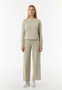 Beżowy sweter z długimi rękawami i prążkowanymi mankietami, w zestawie z szerokimi beżowymi spodniami. Oba elementy wykonane są z miękkiego materiału, zestawione z białymi trampkami.