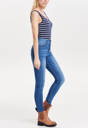 Femme portant un débardeur rayé bleu et blanc, un jean bleu ajusté et des bottines marron, se tenant dans une pose détendue devant un fond uni.