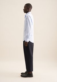 Camicia blu chiaro con bottoni e maniche lunghe, abbinata a pantaloni blu navy e scarpe nere, vista di lato. Tessuto liscio, vestibilità su misura.