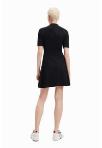 Schwarzes Kurzarmkleid mit schwingendem Saum, gerippter Textur und seitlichen Ausschnitten, kombiniert mit weißen Turnschuhen mit roten Akzenten.