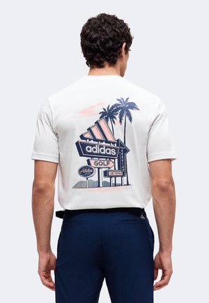 Mann trägt ein weißes T-Shirt mit einem Vintage-Stil adidas Golf-Schild und Palmen-Grafik auf dem Rücken, kombiniert mit dunkelblauer Hose.