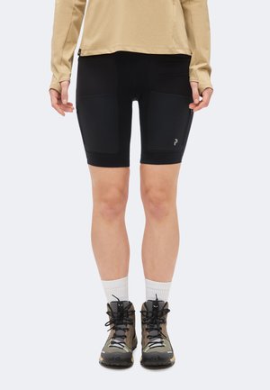Personne portant un haut beige à manches longues, un short de cyclisme noir mi-cuisse avec poches latérales, des chaussettes blanches et des chaussures de randonnée marron, debout.