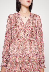 Abito floreale in rosa chiaro con scollatura a V, maniche lunghe e polsini arricciati. Presenta intricate fantasie paisley e floreali con texture sottili.