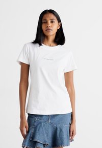 T-shirt bianco in cotone con maniche corte, scollo rotondo e piccolo logo stampato sul petto. Abbinato a una gonna di jeans con orlo arricciato.