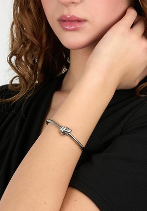 Donna con capelli castani ricci che indossa una maglia nera e un braccialetto d'argento a forma di nodo al polso, mano che tocca il collo.