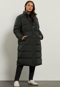 Abrigo acolchado negro largo con un cuello alto, botones de presión y bolsillos laterales. Tiene un diseño acolchado con una textura suave y una longitud hasta la rodilla.