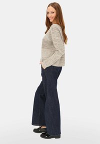 Jasnoszary dzianinowy sweter z długimi rękawami, w połączeniu z szerokimi dżinsami o ciemnym odcieniu i czarnymi butami na platformie. Modelka stoi z jedną ręką w kieszeni.