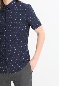 Camicetta a maniche corte, bottonata, di colore blu navy con motivi geometrici bianchi. Presenta un colletto, una tasca frontale e una vestibilità strutturata.