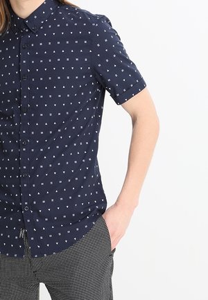 Chemise à manches courtes avec boutons de couleur marine ornée de motifs géométriques blancs. Dotée d'un col, d'une poche avant et d'une coupe structurée.
