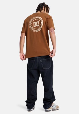 T-shirt en coton marron avec un logo graphique circulaire beige au dos. Associé à un jean en denim foncé et des baskets à motifs.