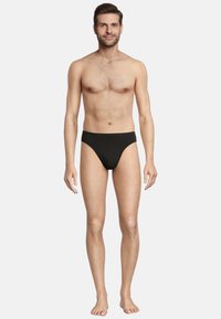 Slip bikini nero realizzato in tessuto morbido, caratterizzato da un design a vita bassa, una cintura elastica e una copertura minima. Il modello è in piedi a piedi nudi.