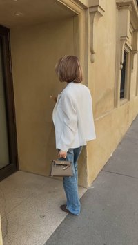 Weißer übergroßer Blazer, hellblaue Jeans und braune Handtasche. Texturierte Wand und Bürgersteig sichtbar, mit einer Tür vorne.