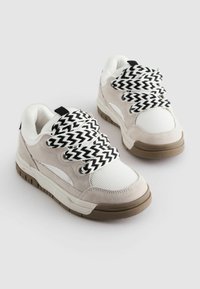 Baskets beiges et blanches avec une semelle solide marron, en daim texturé et en mesh, présentant des lacets à motifs chevrons noirs et blancs.