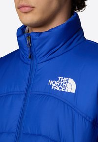 Giacca a vento blu con collo alto, chiusura con zip e logo bianco sul petto. Presenta un tessuto liscio e lucido con una texture trapuntata.