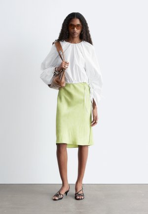 Žena s dlhými kučeravými vlasmi nosí bielu nafúknutú blúzku, svetlozelenú sukňu po kolená, hnedú kabelku cez rameno, slnečné okuliare a čierne sandále na remienky.