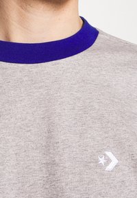 Primer plano de una camisa gris con un cuello morado y un logo de estrella y flecha blanca en el pecho, llevada por una persona.