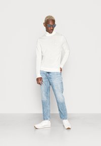 Hvid langærmet turtleneck, lyseblå jeans og hvide sneakers. Tilbehøret inkluderer mørke solbriller. Glat tekstur på toppen.
