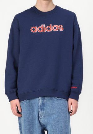 Persona que lleva una sudadera azul marino con un gran logo en rojo y blanco de "adidas" en el pecho y un pequeño logo rojo de "adidas" en el puño de la manga izquierda.