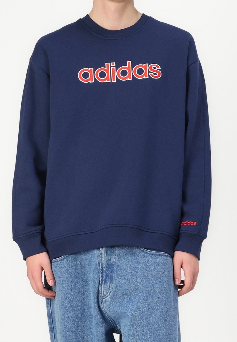 Person som bär en marinblå sweatshirt med en stor röd och vit "adidas"-logotyp på bröstet och en liten röd "adidas" på cuffs på vänster ärm.