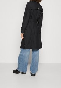 Svart trench coat med bälte i midjan, med en ventil bak, axelkläppar och långa ärmar, i kombination med vida blå jeans.