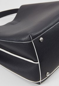 Coin d'un sac à main en cuir noir texturé avec un liseré blanc et des clous en métal argenté sur fond blanc.
