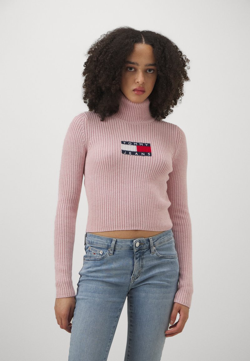 Maglione a collo alto a coste rosa chiaro con maniche lunghe, caratterizzato da un logo "TOMMY JEANS" ricamato su una toppa blu. Indossato con jeans blu.