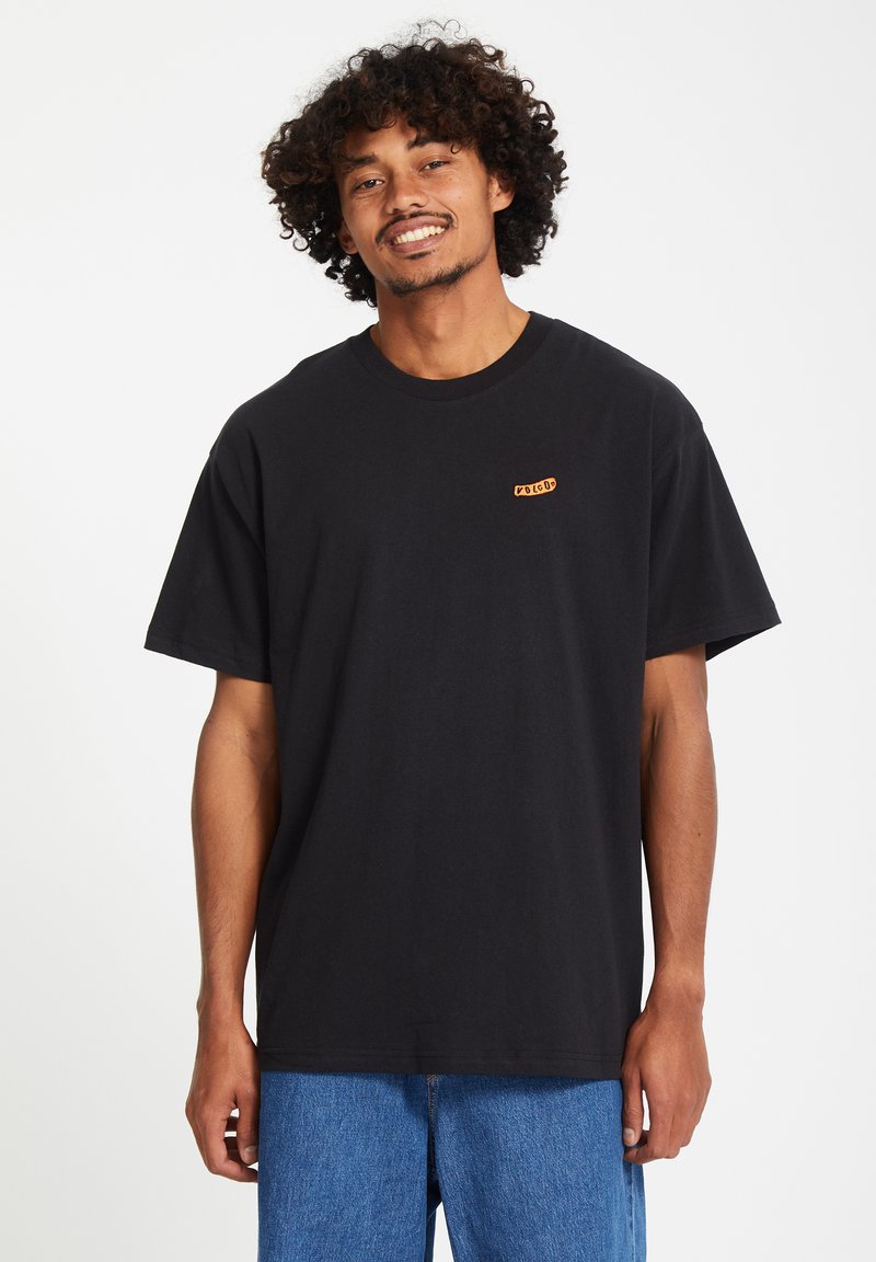 Zwarte katoenen t-shirt met korte mouwen en ronde hals; heeft een klein oranje logo op de linker borst. Draagt bij blauwe denimbroekjes.