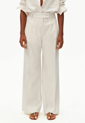 Donkergekleurde persoon die beige, hooggetailleerde, wijde broek draagt, een wit overhemd met knopen en opgerolde mouwen en sandalen met een met kralen versierde teent ring.