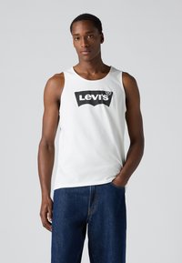 Bijela pamučna majica bez rukava s crnim Levi's logom. Model nosi plave traperice. Jednostavan dizajn, bez rukava s okruglim izrezom.