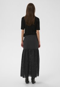 Schwarzes, geripptes Kurzarmoberteil kombiniert mit einem schwarzen Maxirock, der eine spitzenähnliche Überlagerung und einen gewellten Saum aufweist. Glatte Textur mit mehrlagigem Design.