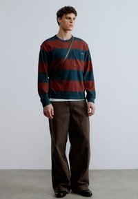 Μπλούζα με μακριά μανίκια και ρίγες σε σκούρο πράσινο και μπορντό, με τσέπη στο στήθος, συνδυασμένη με καφέ παντελόνι φαρδύ. Φοριούνται μαύρα παπούτσια.