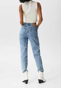Jeans taille haute bleu clair avec une finition texturée, dotés de poches arrière et d'un patch en cuir beige de marque. Portés avec un haut sans manches et des bottines blanches.