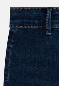 Temno modra denim tkanina v bližnjem načinu, ki ima visok pas z vidnimi šivi in zanko za pas. Surovina je gladka s blagim sijajem.