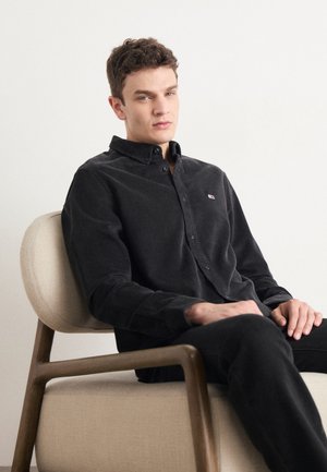 Camicia nera con bottoni realizzata in un tessuto testurizzato, con un piccolo logo sul petto, abbinata a pantaloni neri, indossata da un modello maschile seduto.