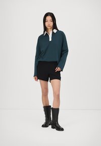 Temno teal dolgi rokav polo majica z belim ovratnikom, v kombinaciji s črnimi fitted kratkimi hlačami in črnimi visokimi škornji do kolen. Minimalistična zasnova.