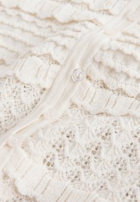 Vêtement tricoté de couleur crème avec des couches à volants, des motifs en dentelle et de petits boutons ronds crème le long de l’ouverture avant.