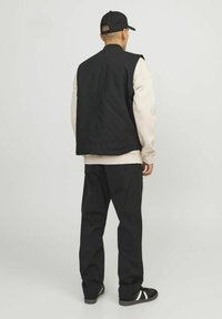 Sort ærmeløs vest over en beige sweatshirt, parret med sorte bukser og sorte sko med hvide striber. Enkel design, sporty look.